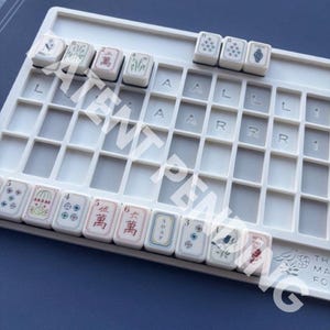May include: A white plastic Mahjong tile rack with a grid of empty slots and several rectangular tiles placed along the top and bottom edges. The tiles feature various colorful illustrations, Chinese characters, and the word SOAP printed in black ink.