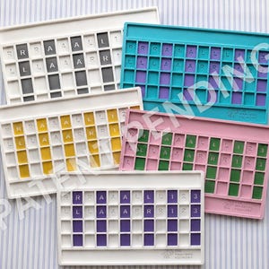 Op de afbeelding: Vijf rechthoekige mahjong-tegelhouders in wit, taling, geel, roze en paars. Elke houder heeft een raster met de letters R, L, A en de cijfers 1, 2, 3 op de tegels. De merknaam The Mahjong Foundry is op elk exemplaar in reli&euml;f aangebracht.
