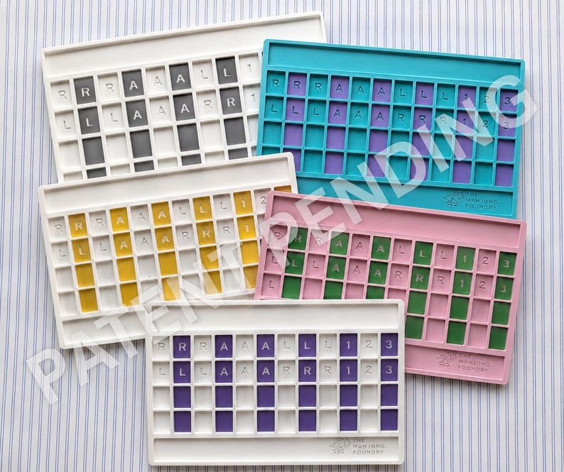 Op de afbeelding: Vijf rechthoekige mahjong-tegelhouders in wit, taling, geel, roze en paars. Elke houder heeft een raster met de letters R, L, A en de cijfers 1, 2, 3 op de tegels. De merknaam The Mahjong Foundry is op elk exemplaar in reli&euml;f aangebracht.