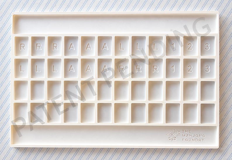 Op de afbeelding: Een rechthoekig mahjong-tegelrek van gebroken wit plastic met een raster van achtenveertig verzonken vakjes. De bovenste twee rijen bevatten in reli&euml;f gedrukte letters R, A, L en de cijfers 1, 2, 3. Rechtsonder staat het logo van The Mahjong Foundry.