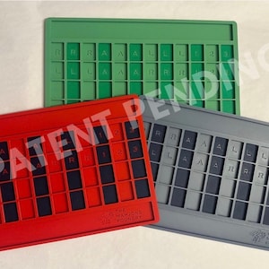 Op de afbeelding: Drie rechthoekige siliconen mahjong-tegelhouders in groen, rood en grijs. Elke houder heeft een raster met sleuven voor tegels, met de letters R, A, L en de cijfers 1, 2, 3 in reli&euml;f op het oppervlak. De rode en grijze houders tonen het logo.