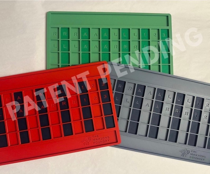 Op de afbeelding: Drie rechthoekige siliconen mahjong-tegelhouders in groen, rood en grijs. Elke houder heeft een raster met sleuven voor tegels, met de letters R, A, L en de cijfers 1, 2, 3 in reli&euml;f op het oppervlak. De rode en grijze houders tonen het logo.