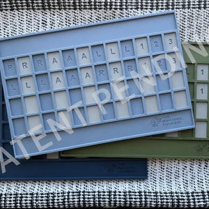 Op de afbeelding: Drie rechthoekige mahjong-tegelhouders in marineblauw, lichtblauw en olijfgroen. Elke houder heeft een raster met sleuven voor tegels, waarbij sommige sleuven de letters R, A, L en de cijfers 1, 2, 3 bevatten. Het Mahjong Foundry-logo is in elk exemplaar gegraveerd.