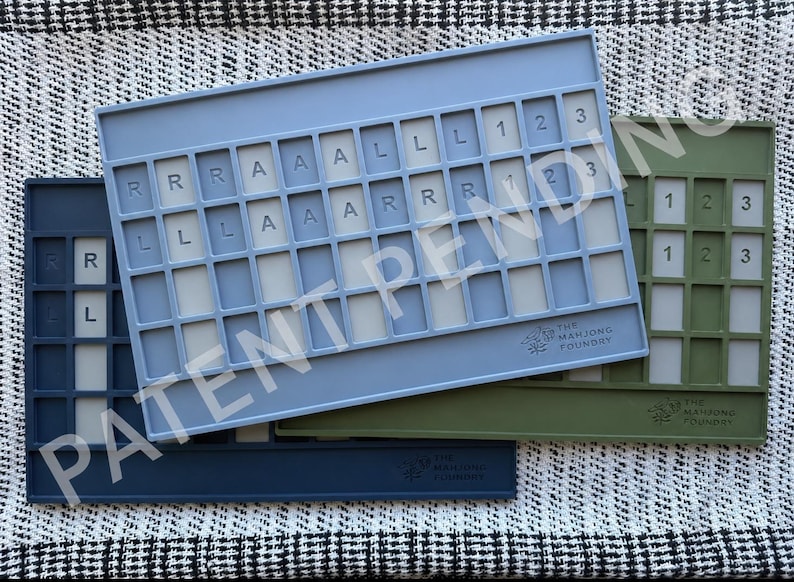 Op de afbeelding: Drie rechthoekige mahjong-tegelhouders in marineblauw, lichtblauw en olijfgroen. Elke houder heeft een raster met sleuven voor tegels, waarbij sommige sleuven de letters R, A, L en de cijfers 1, 2, 3 bevatten. Het Mahjong Foundry-logo is in elk exemplaar gegraveerd.