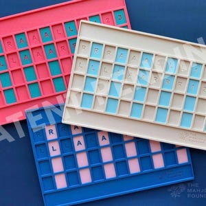 Op de afbeelding: Drie rechthoekige siliconen mahjong-tegelhouders in roze, beige en blauw. Elke houder heeft een raster van verzonken sleuven met in reli&euml;f aangebrachte letters R, L, A en cijfers 1, 2, 3. De beige en blauwe houders tonen het logo The Mahjong Foundry.