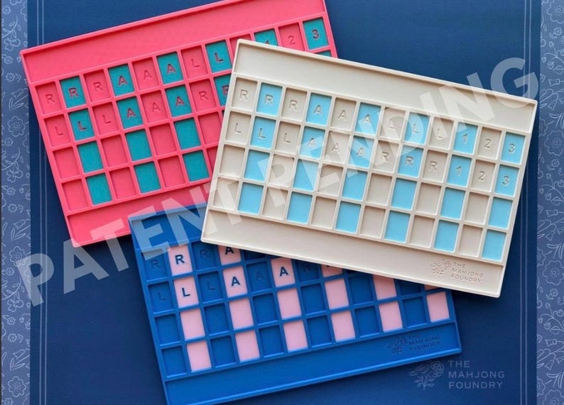Op de afbeelding: Drie rechthoekige siliconen mahjong-tegelhouders in roze, beige en blauw. Elke houder heeft een raster van verzonken sleuven met in reli&euml;f aangebrachte letters R, L, A en cijfers 1, 2, 3. De beige en blauwe houders tonen het logo The Mahjong Foundry.