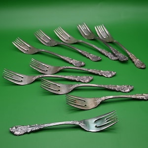 May include: A collection of antique silver forks with ornate floral detailing on the handles. The forks are arranged on a solid green background, showcasing their tines and decorative elements. The silverware is likely for formal dining.