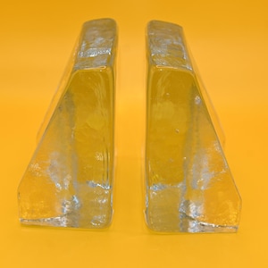 May include: Two clear glass bookends with a textured, frosted appearance. The bookends are wedge-shaped and stand upright against a bright yellow background. The glass has a slightly yellow tint.
