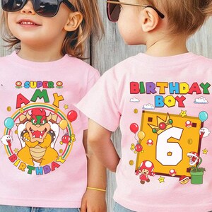May include: Pink t-shirts with colorful Super Mario-themed birthday graphics. One shirt says "Super Amy Birthday" with Bowser, balloons, and a rainbow. The other says "Birthday Boy 6" with Mario characters and a gold frame.