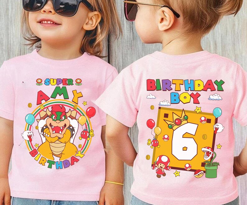May include: Pink t-shirts with colorful Super Mario-themed birthday graphics. One shirt says "Super Amy Birthday" with Bowser, balloons, and a rainbow. The other says "Birthday Boy 6" with Mario characters and a gold frame.