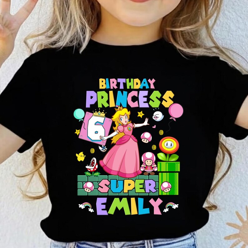 Op de afbeelding: Zwart T-shirt met een kleurrijk grafisch ontwerp. Het ontwerp bevat de tekst "BIRTHDAY PRINCESS", het nummer "6" en de naam "EMILY". De afbeelding bevat ook stripfiguren en elementen uit een videogame.