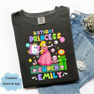 Op de afbeelding: Donkergrijs t-shirt met een kleurrijke afbeelding van Princess Peach, de tekst "BIRTHDAY PRINCESS 6 SUPER EMILY" en andere Super Mario Bros. karakters. Het shirt wordt getoond met een blauwe jeans.