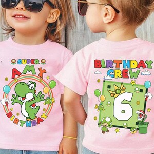 May include: Pink t-shirts with colorful graphics. One shirt features a green Yoshi character and the text "SUPER AMY BIRTHDAY". The other shirt displays "BIRTHDAY CREW" and a number 6 design with cartoon characters.