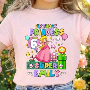 Op de afbeelding: Roze T-shirt met een kleurrijke afbeelding van Princess Peach uit Super Mario, met de tekst "BIRTHDAY PRINCESS 6 SUPER EMILY". Het ontwerp bevat ballonnen, munten en andere game-elementen.