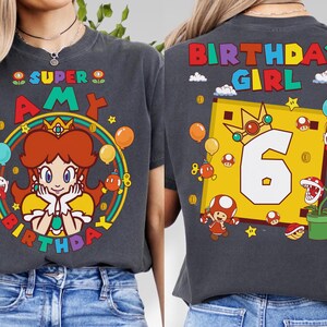 May include: Dark gray t-shirt featuring a colorful graphic of a cartoon princess with the text "SUPER AMY BIRTHDAY" on the front. The back of the shirt has "BIRTHDAY GIRL" and the number "6". Balloons and stars are also featured.