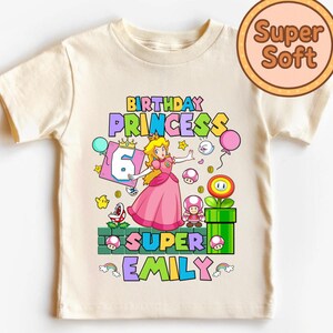 Op de afbeelding: Een cr&egrave;mekleurig T-shirt met een kleurrijke afbeelding van Princess Peach uit Super Mario. Het ontwerp bevat de tekst "Birthday Princess", het nummer "6" en de naam "Emily". Een "Super Soft" logo staat rechtsboven.