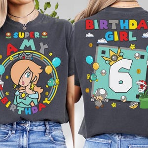 May include: Dark gray t-shirt featuring a colorful Super Mario-themed design. The front displays "Super Amy" with a character illustration, balloons, and stars. The back shows "Birthday Girl" with the number "6" and more character graphics.