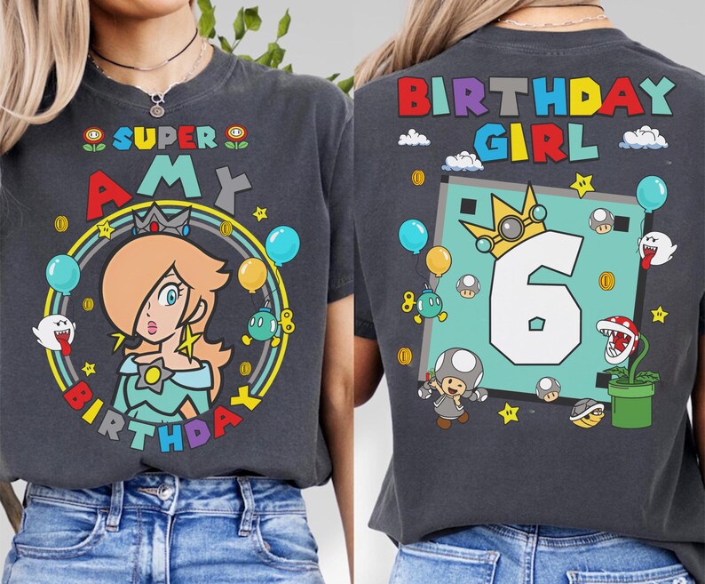 May include: Dark gray t-shirt featuring a colorful Super Mario-themed design. The front displays "Super Amy" with a character illustration, balloons, and stars. The back shows "Birthday Girl" with the number "6" and more character graphics.