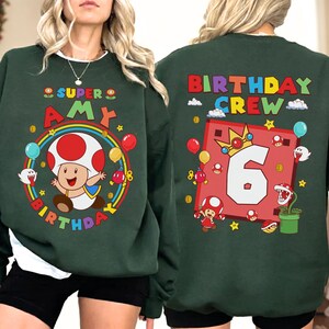 May include: Dark green sweatshirt featuring a colorful graphic of a mushroom character, a rainbow, and the text "SUPER AMY BIRTHDAY" on the front. The back of the sweatshirt has the text "BIRTHDAY CREW" and a large number "6".