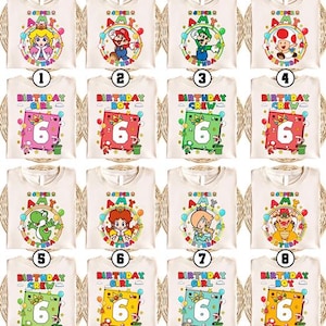 May include: A collection of cream-colored t-shirts featuring various cartoon characters and birthday-themed designs. Each shirt has a different character, such as Mario, Luigi, or Princess Peach, with the text "Super Amy" and a number 6. Some shirts say "Birthday Girl," "Birthday Boy," or "Birthday Crew."