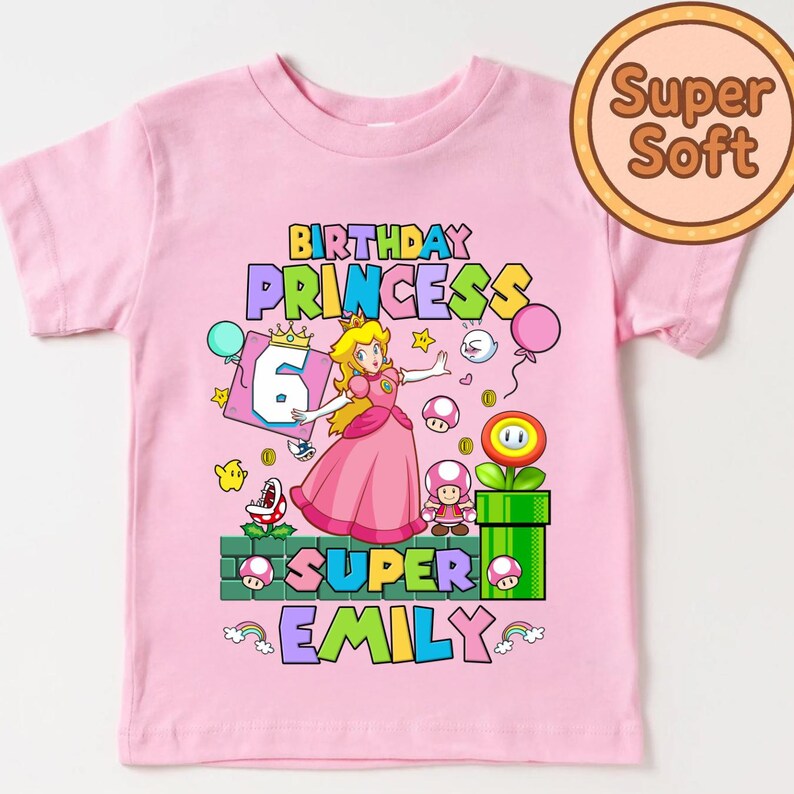 Op de afbeelding: Roze t-shirt met een kleurrijke afbeelding van Princess Peach uit de Super Mario-videogameserie. Het ontwerp bevat de tekst "Birthday Princess 6 Super Emily" met game-gerelateerde elementen.