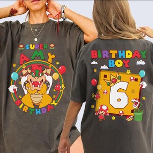 May include: Dark gray t-shirts with colorful Super Mario-themed graphics. One shirt says "Super Amy Birthday" with a Bowser character. The other says "Birthday Boy 6" with Mario characters and a crown. Balloons and stars decorate the designs.