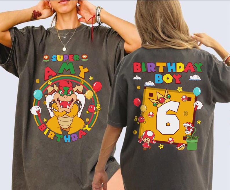 May include: Dark gray t-shirts with colorful Super Mario-themed graphics. One shirt says "Super Amy Birthday" with a Bowser character. The other says "Birthday Boy 6" with Mario characters and a crown. Balloons and stars decorate the designs.