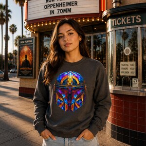 K&ouml;nnte beinhalten: Ein anthrazitfarbenes Langarm-Sweatshirt mit einer leuchtenden, mehrfarbigen Grafik eines Helms mit geometrischen Mustern in Blau-, Lila-, Orange- und Gelbt&ouml;nen, kombiniert mit einer hellblauen Jeanshose aus Denim.