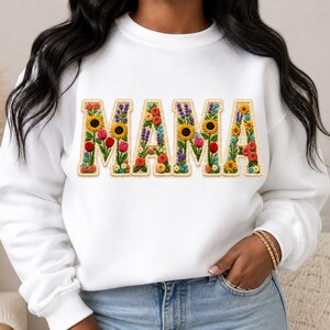 Może przedstawiać: Biała bluza z napisem "MAMA" wyszytym kwiatowym wzorem. Litery wypełnione są kolorowymi kwiatami, w tym słonecznikami, tulipanami i lawendą. Bluza ma okrągły dekolt i długie rękawy.