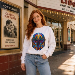 K&ouml;nnte beinhalten: Ein wei&szlig;es Langarm-Sweatshirt mit einer farbenfrohen Grafik im Buntglas-Stil eines Helms auf der Vorderseite. Das Design kombiniert leuchtende Blau-, Orange- und Gelbt&ouml;ne in einem geometrischen Muster, kombiniert mit einer hellblauen Jeanshose.