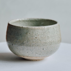 以下が含まれることがあります： 淡い緑色の斑点がある釉薬が施された丸い陶器のボウルで、表面には細かいひび割れ模様があります。ボウルは口が広く、底に向かって細くなっており、釉薬のかかっていない素焼きの茶色の土肌が見える自然な風合いのデザインです。