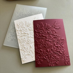 Peut inclure: Un dossier de gaufrage rectangulaire en plastique translucide présentant un motif floral et papillon en relief, accompagné d'une carte blanche et d'une carte rouge foncé, toutes deux gaufrées avec le même motif détaillé de fleurs.