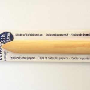 May include: A paper tool made of solid bamboo, 5.8 inches long. The tool is light brown and has a rounded handle with a pointed end. The packaging reads "Fold and score papers."