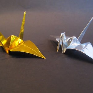 May include: Two origami cranes, one gold and one silver, are displayed against a dark background. The gold crane is on the left, and the silver crane is on the right. Both cranes are folded from metallic paper, showcasing the art of origami.