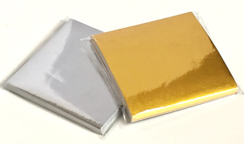 May include: Two stacks of square, metallic paper sheets, one silver and one gold, are shown in a clear plastic wrapper. The paper has a glossy finish, reflecting light. The gold paper has a warm, rich tone, while the silver paper is bright.
