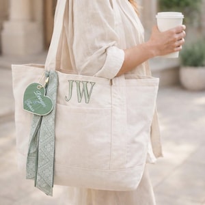 Puede incluir: Bolso tote de lona color crema con las iniciales bordadas en verde "JW" y una etiqueta en forma de corazón. Una bufanda estampada verde claro y una etiqueta verde en forma de corazón están unidas al bolso.