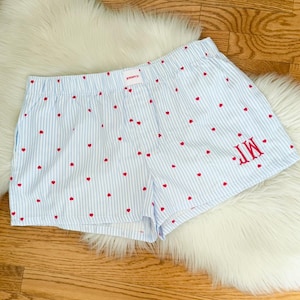 May include: Light blue and white striped shorts with red heart patterns. The shorts have an elastic waistband and a monogram with the initials "M.T." in red. The shorts are made of a lightweight fabric.