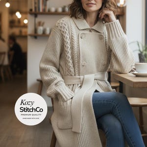 May include: A cream cable-knit cardigan with a double-breasted design and a matching belt. The longline sweater features a shawl collar and ribbed cuffs. The "Kozy StitchCo" logo is visible in the lower left corner. The person is wearing blue jeans.