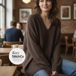 Könnte beinhalten: Eine Frau trägt einen braunen V-Ausschnitt-Pullover und blaue Jeans. Der Pullover hat lange Ärmel und eine lockere Passform. Das Markenlogo "Kozy StitchCo" ist in einem weißen Kreis sichtbar. Der Hintergrund zeigt ein Café.