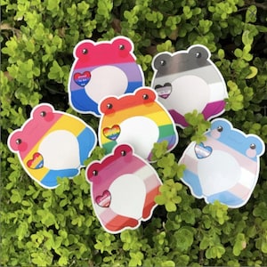 May include: A collection of six frog-shaped stickers with various LGBTQ+ pride flag designs. Each sticker features a different flag, including bisexual, pansexual, lesbian, transgender, asexual, and rainbow pride flags. The stickers are set against a backdrop of green foliage.