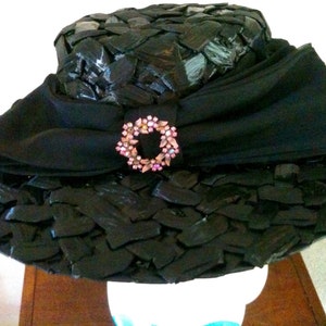 May include: A black hat with a wide brim and a decorative bow. The hat features a textured design and a circular embellishment with pink and clear stones. The hat is likely a vintage or retro style.