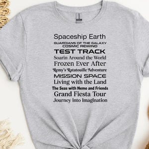 Op de afbeelding: Een lichtgrijs t-shirt met zwarte tekst met attracties van een themapark, waaronder "Spaceship Earth" en "Test Track". De shirt is aan de onderkant geknoopt. Een paarse cirkel met "Sports Grey" staat linksonder.