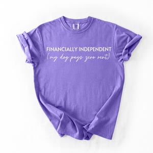 Könnte beinhalten: Ein violettes Kurzarm-T-Shirt mit dem weißen Text FINANCIALLY INDEPENDENT und dem kursiven Text (my dog pays zero rent) auf der Brust. Das Kleidungsstück hat einen Rundhalsausschnitt und eine lockere Passform mit hochgekrempelten Ärmeln.