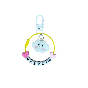 May include: A keychain with a light blue clasp and a yellow wire loop. The loop holds a white cloud charm with a smiling face, pink hearts, and a beaded word "KEYZY".