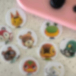 May include: A collection of round, white stickers featuring colorful cartoon animal characters. The stickers are arranged on a light-colored surface, with a pink electronic device in the background.