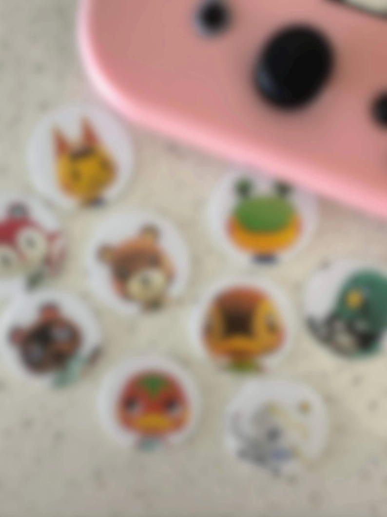 May include: A collection of round, white stickers featuring colorful cartoon animal characters. The stickers are arranged on a light-colored surface, with a pink electronic device in the background.