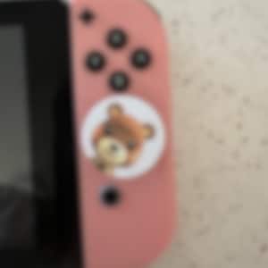 May include: A pink Nintendo Switch Joy-Con controller with black buttons and a circular sticker featuring a cartoon bear. The controller is partially visible, showing the buttons and the attached sticker.
