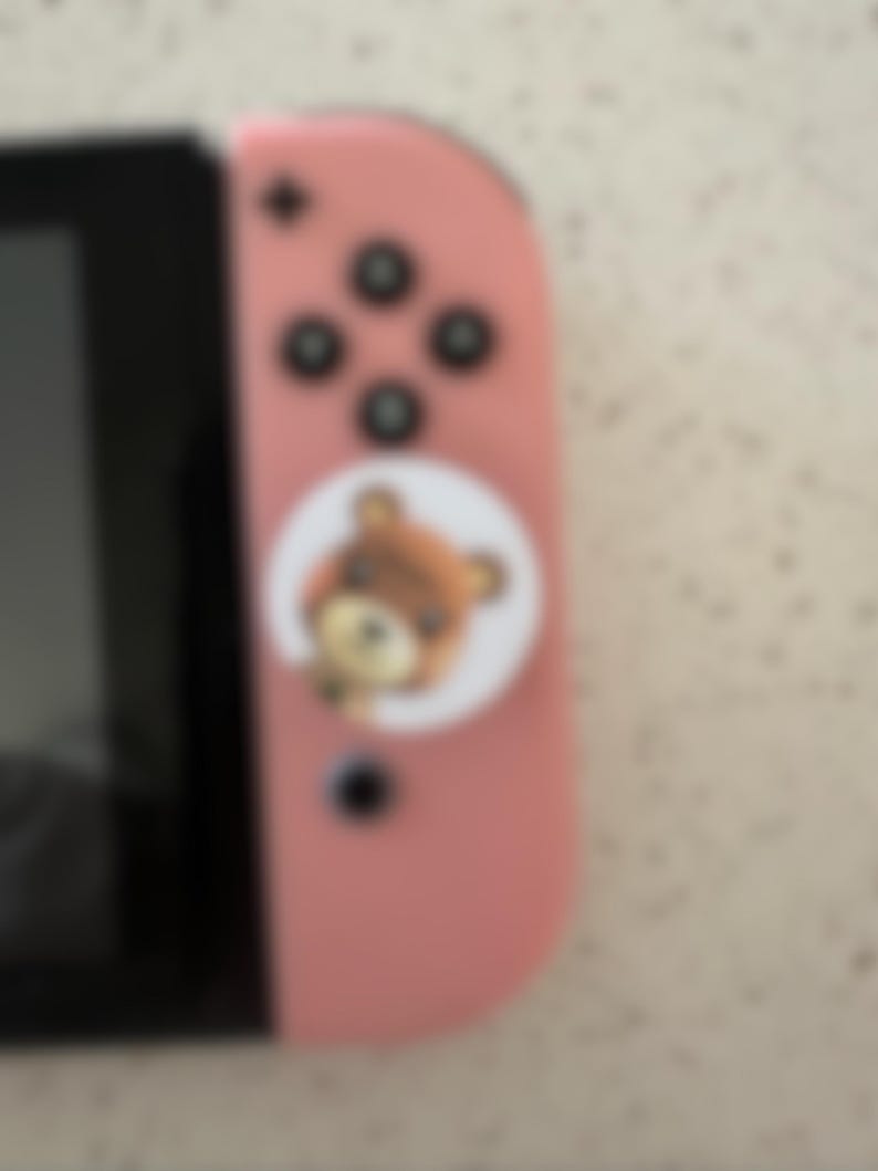 May include: A pink Nintendo Switch Joy-Con controller with black buttons and a circular sticker featuring a cartoon bear. The controller is partially visible, showing the buttons and the attached sticker.