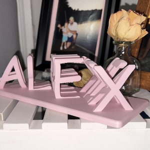 Puede incluir: Un letrero de nombre de madera rosa que deletrea "ALEX". Las letras están conectadas y montadas sobre una base rectangular. El letrero se muestra sobre una superficie blanca, con fotos enmarcadas y un jarrón con una flor seca en el fondo.