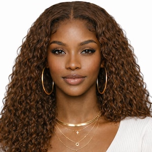 May include: A woman with brown curly hair, wearing large gold hoop earrings and multiple gold necklaces. The necklaces include a choker, a cross pendant, and a circle pendant. She is wearing a white top.